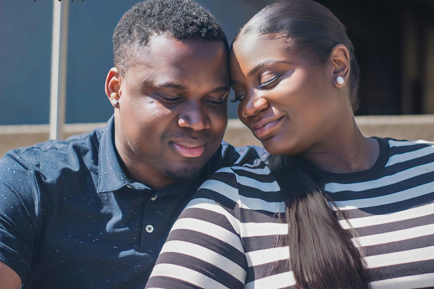 A loving couple leaning heads together, smiling warmly, man in blue polo and woman in striped top, outdoors in sunlight.
