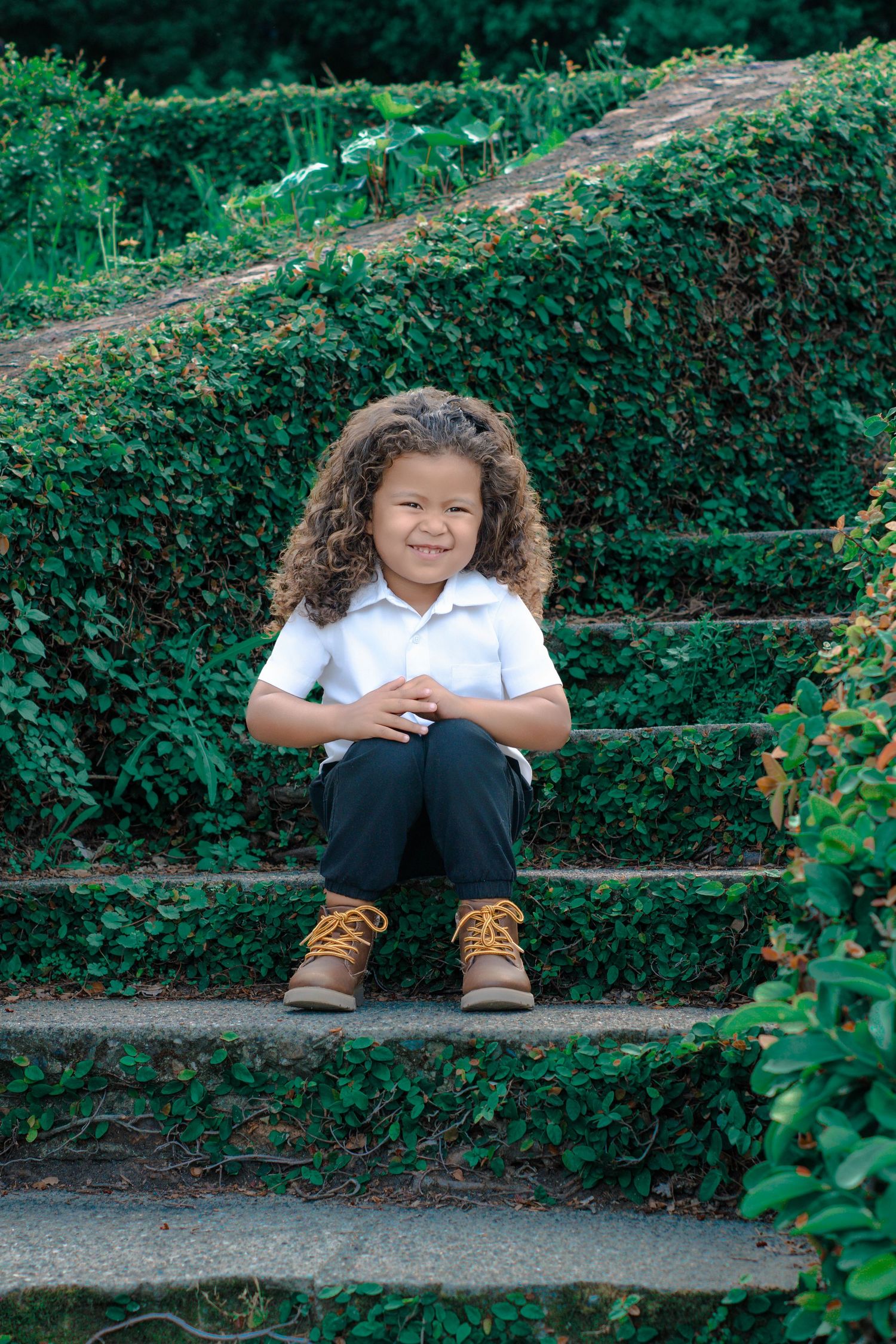 Smiling child with curly hair sitting on stone steps surrounded by lush green hedges, wearing white polo and brown boots.
