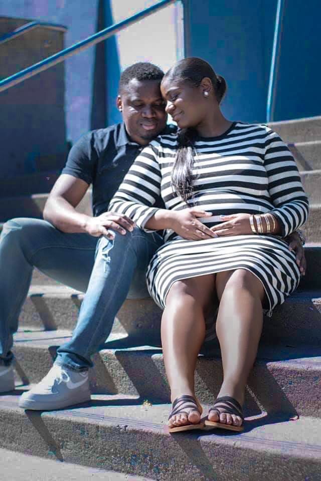 A pregnant couple sits on outdoor steps, the woman in a striped dress cradling her belly while her partner holds her hand.