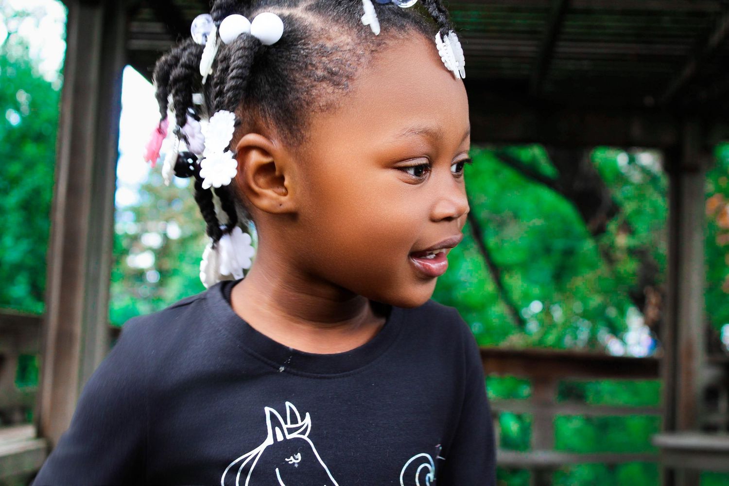 A cute young Black girl with braided pigtails and white bows wears a black horse-print shirt in an outdoor setting.
