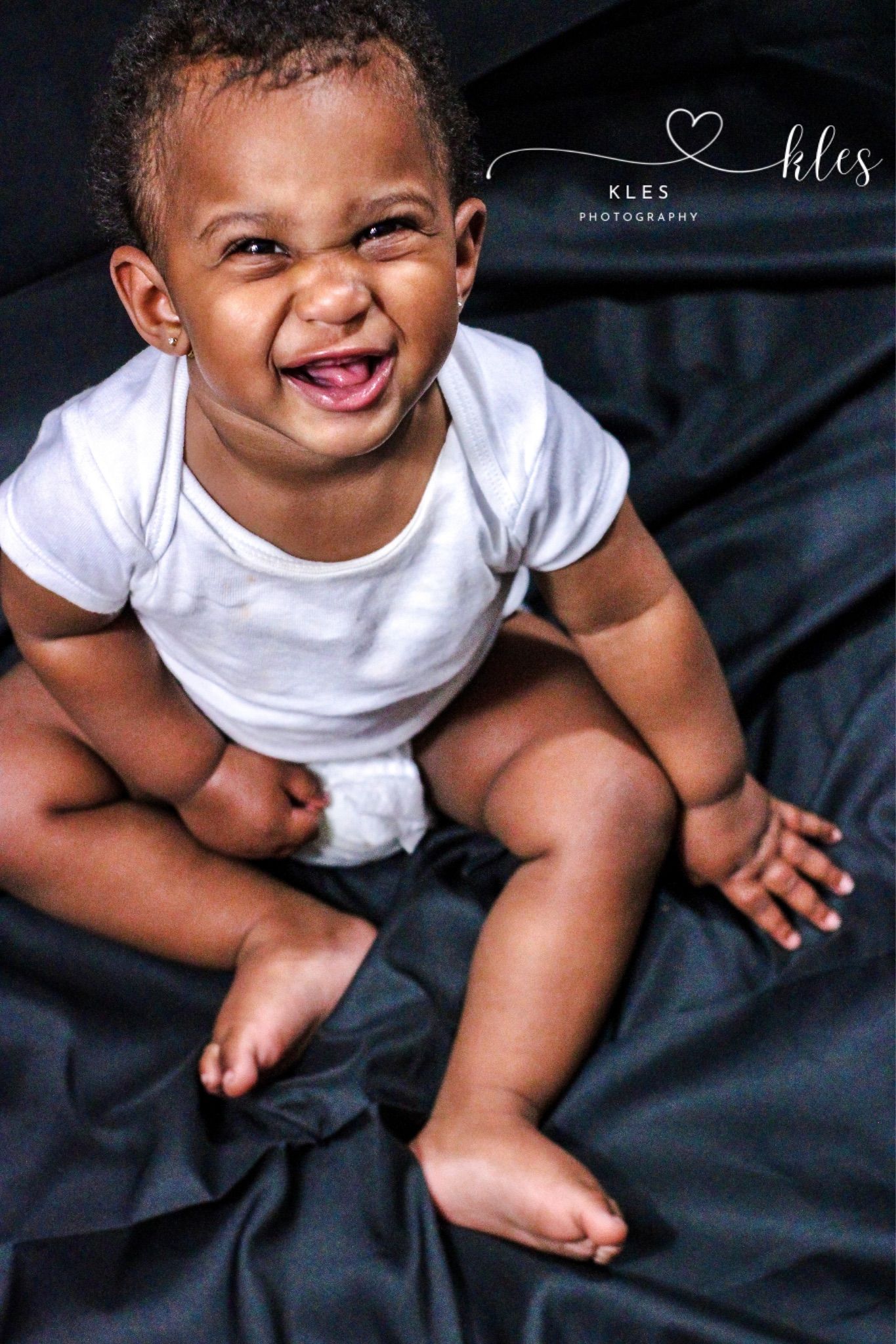 Adorable laughing baby in white onesie sitting on dark fabric, photographed by Kles Photography.