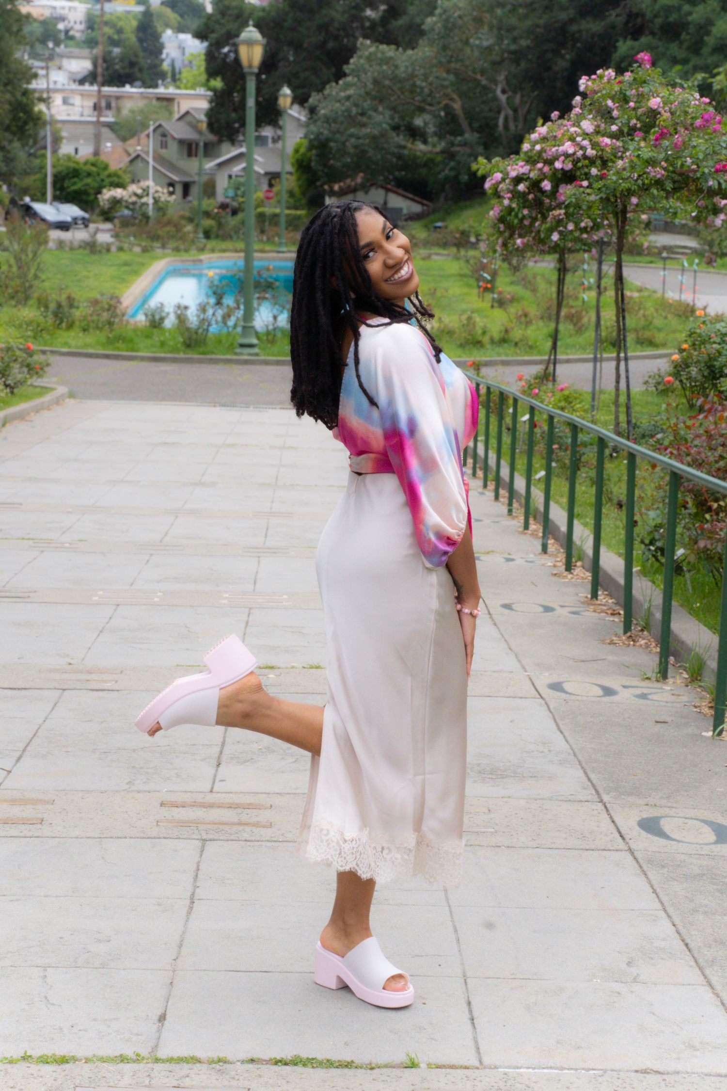 Joyful Black woman in flowing white dress with colorful wrap laughing on garden steps wearing pink heels.