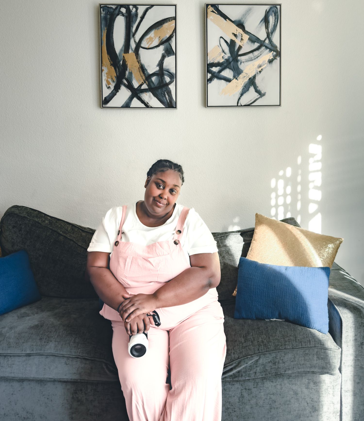 Woman in pink overalls sitting on gray sofa with blue and gold pillows beneath two abstract black and gold wall art pieces.