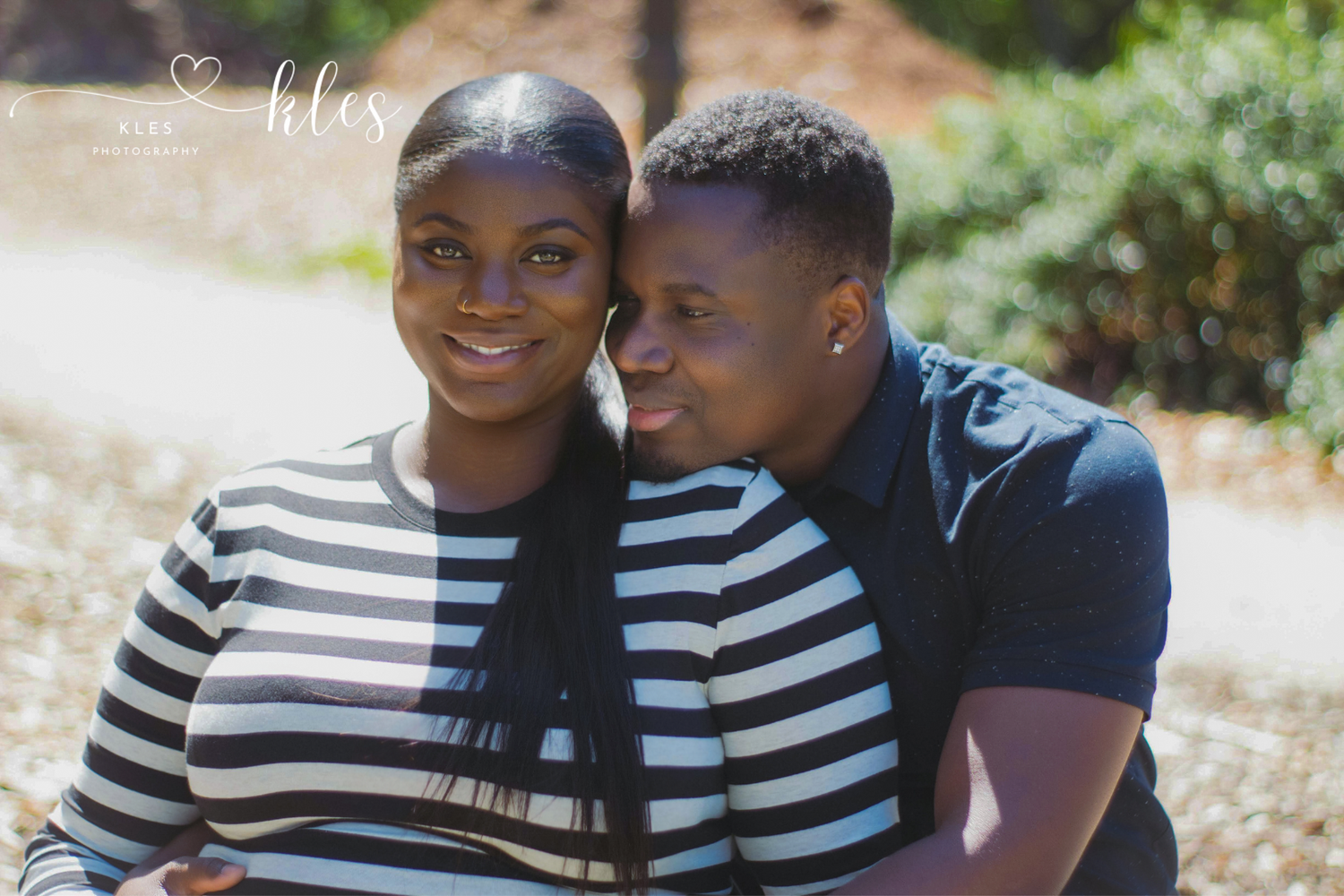 A smiling couple in matching striped shirts poses outdoors in a park for a professional portrait photo.