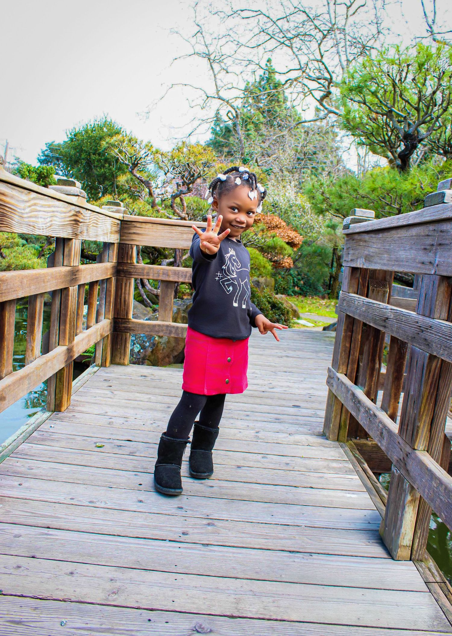 Young girl in red skirt and black top makes peace sign on wooden bridge in a lush garden park.