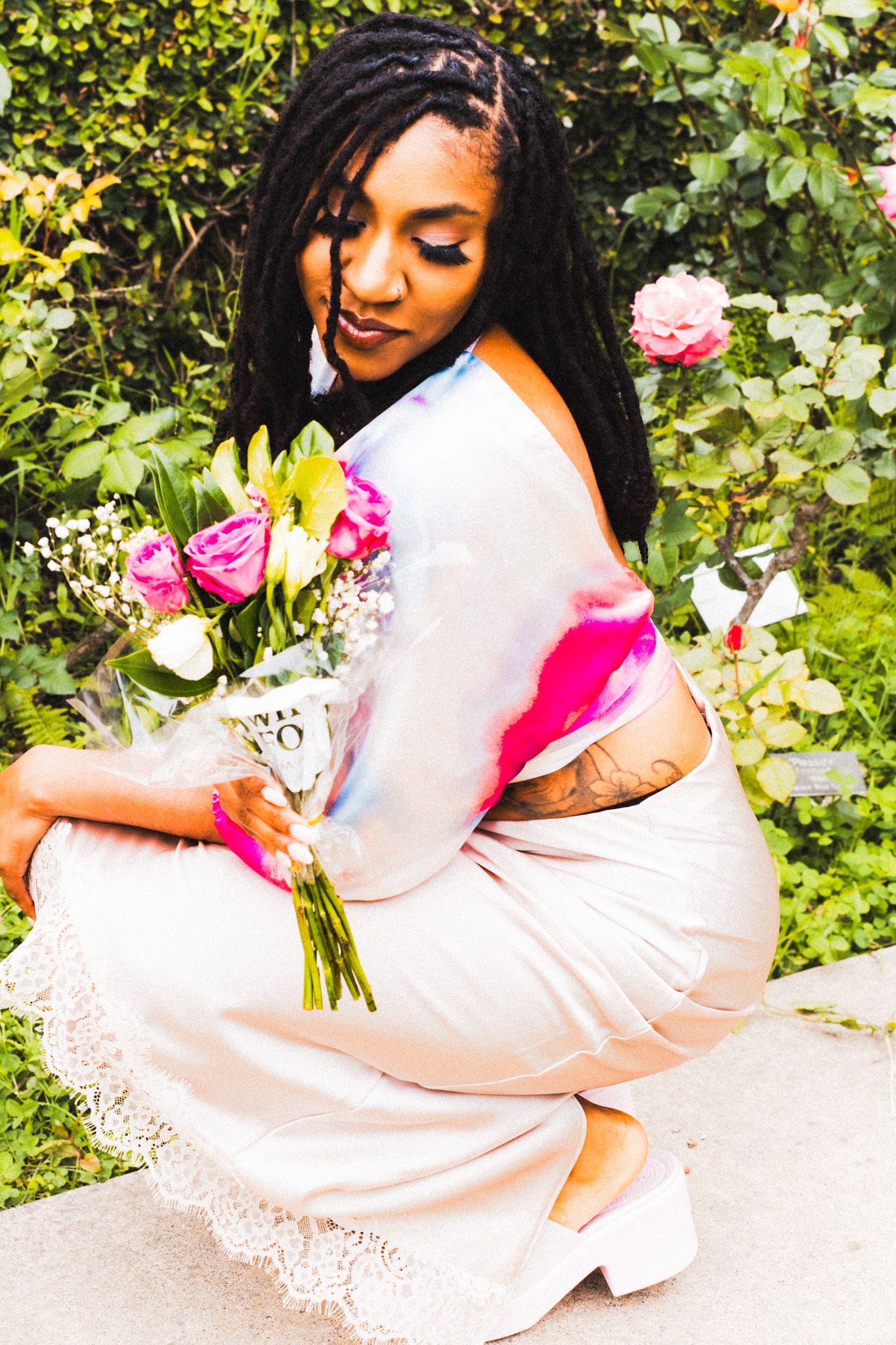 Woman in colorful tie-dye top and cream lace skirt crouching with pink roses bouquet in garden.