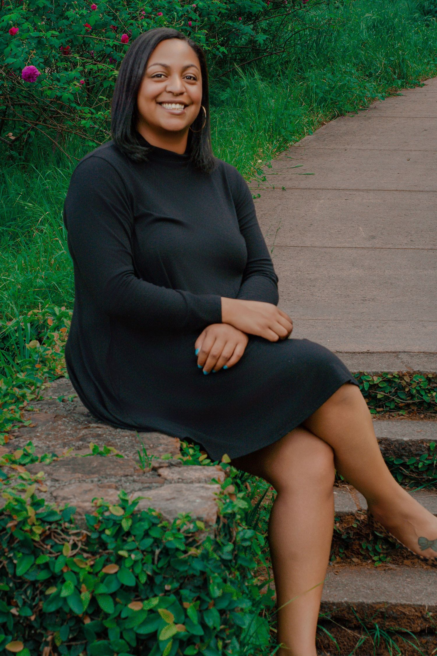A smiling Black woman in a dark green long-sleeve dress sits on stone steps surrounded by lush greenery.