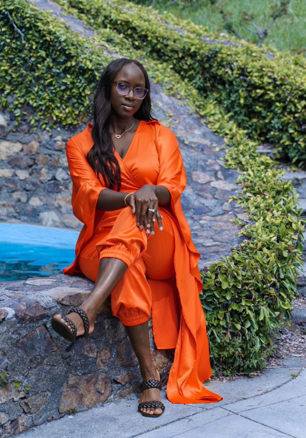 Fashionable Black woman in bold orange satin jumpsuit and duster sitting on stone wall in garden.