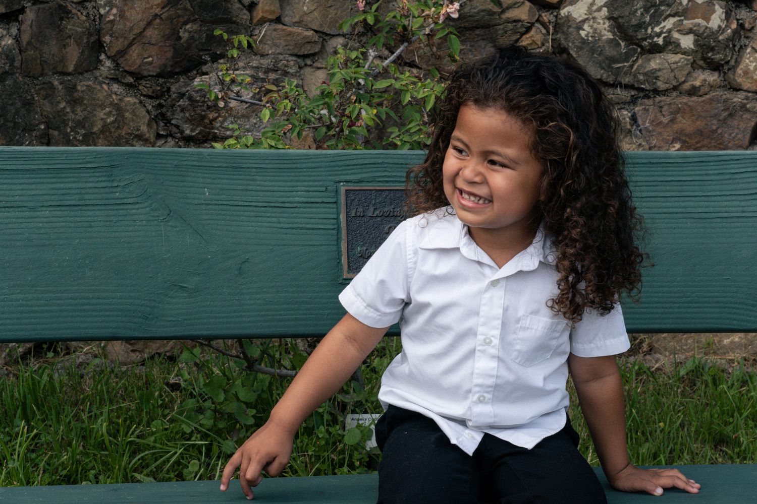 Curly-haired child in a white shirt sitting on a green bench outdoors smiling to the side.