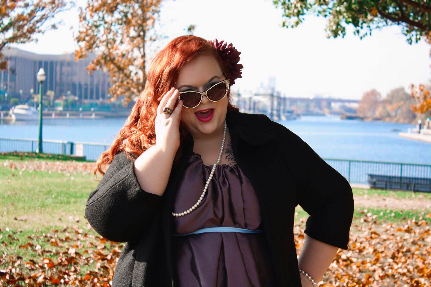 Red-haired woman in vintage sunglasses, pearl necklace, and black cardigan posing outdoors near a river in autumn.