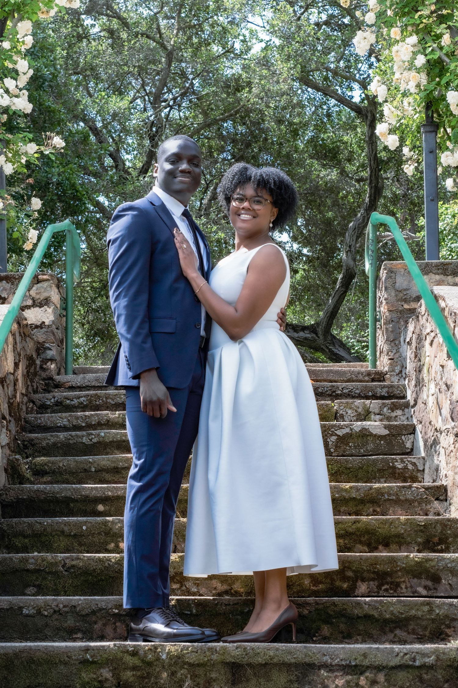 A well-dressed Black couple poses on stone steps surrounded by blooming roses, the woman wearing a white midi dress.