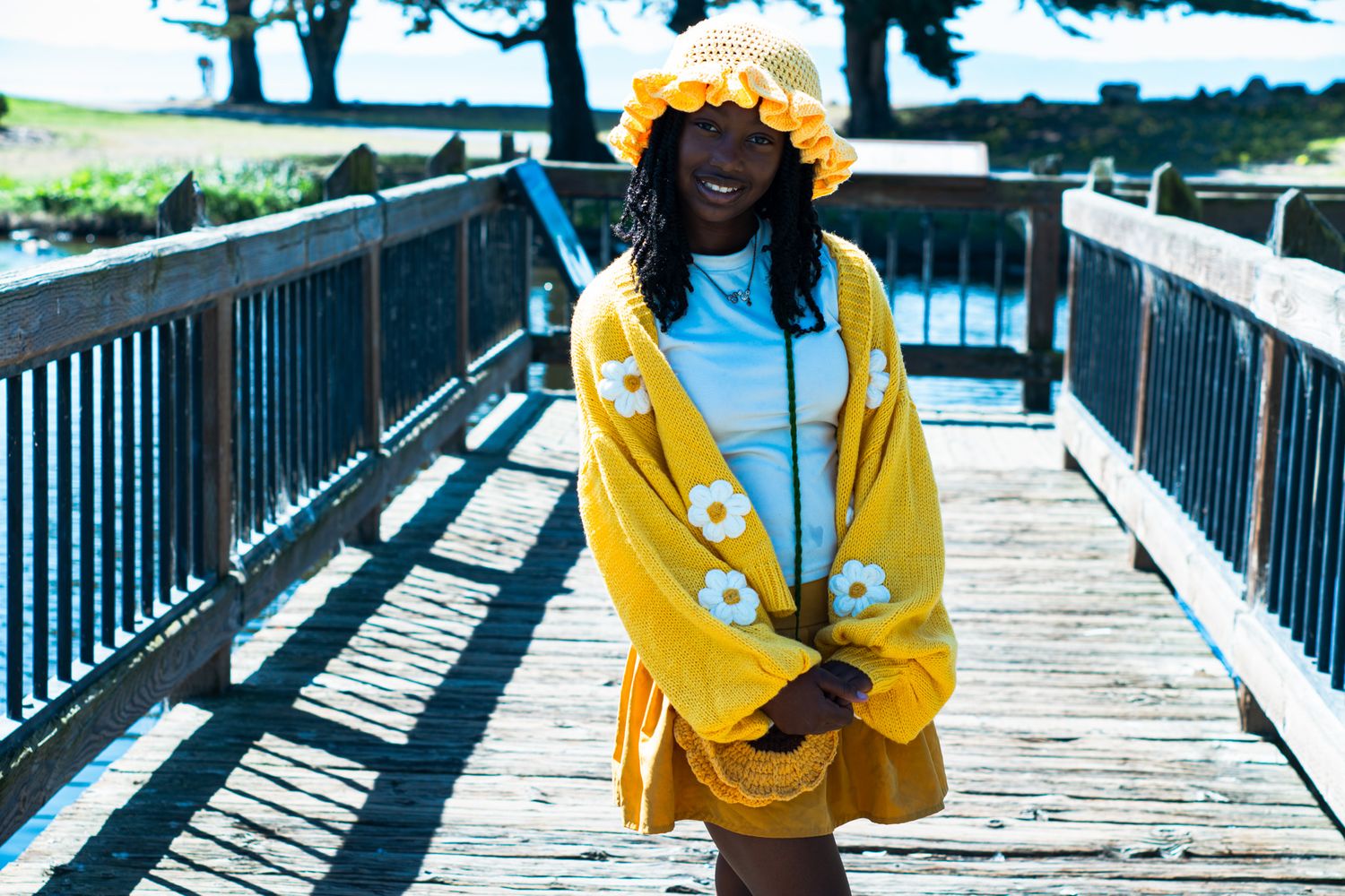 Happy young woman in yellow floral outfit and bucket hat standing on a sunny wooden boardwalk.