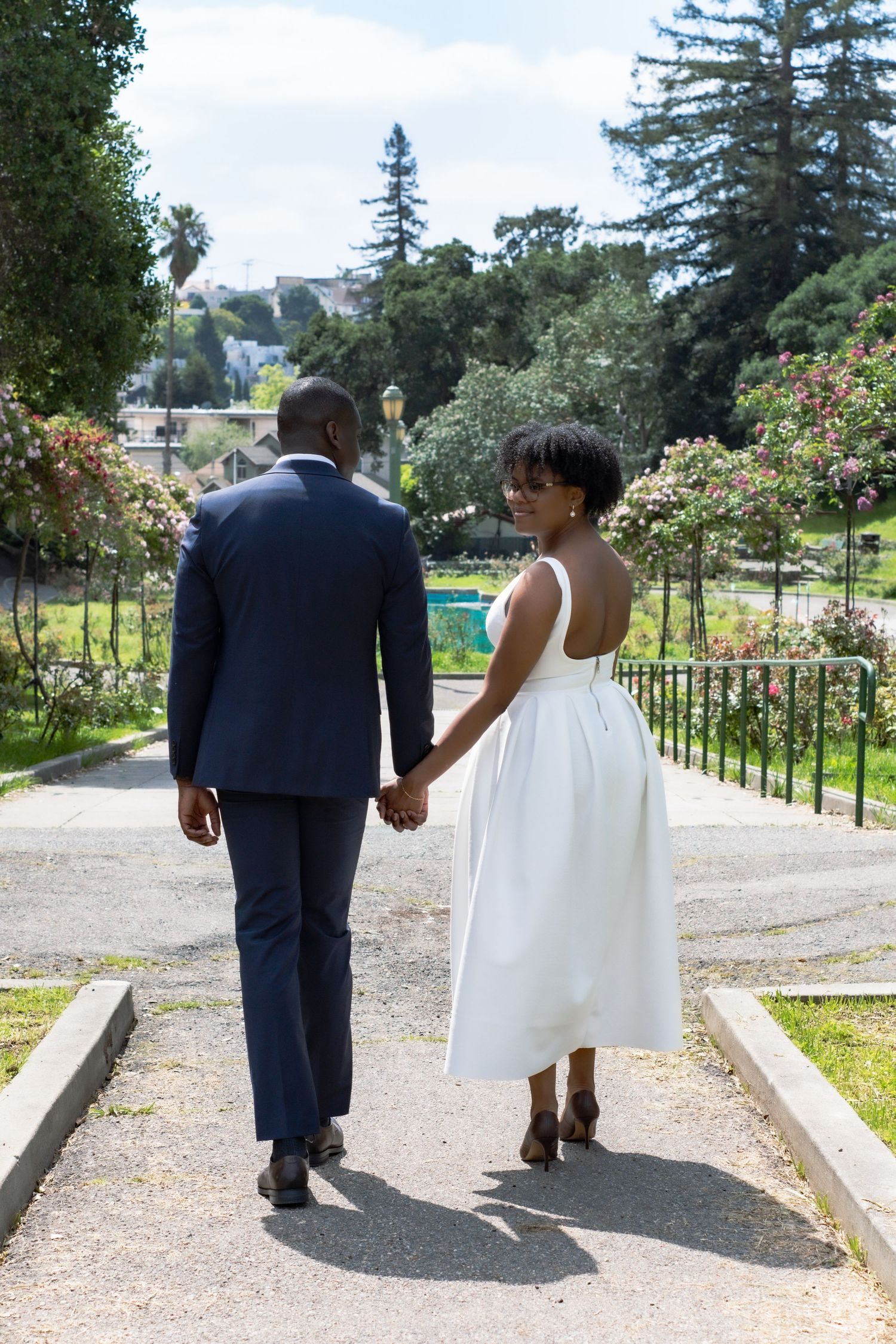 A Black couple in wedding attire holds hands while walking through a lush garden park, the bride glancing back over her shoulder.