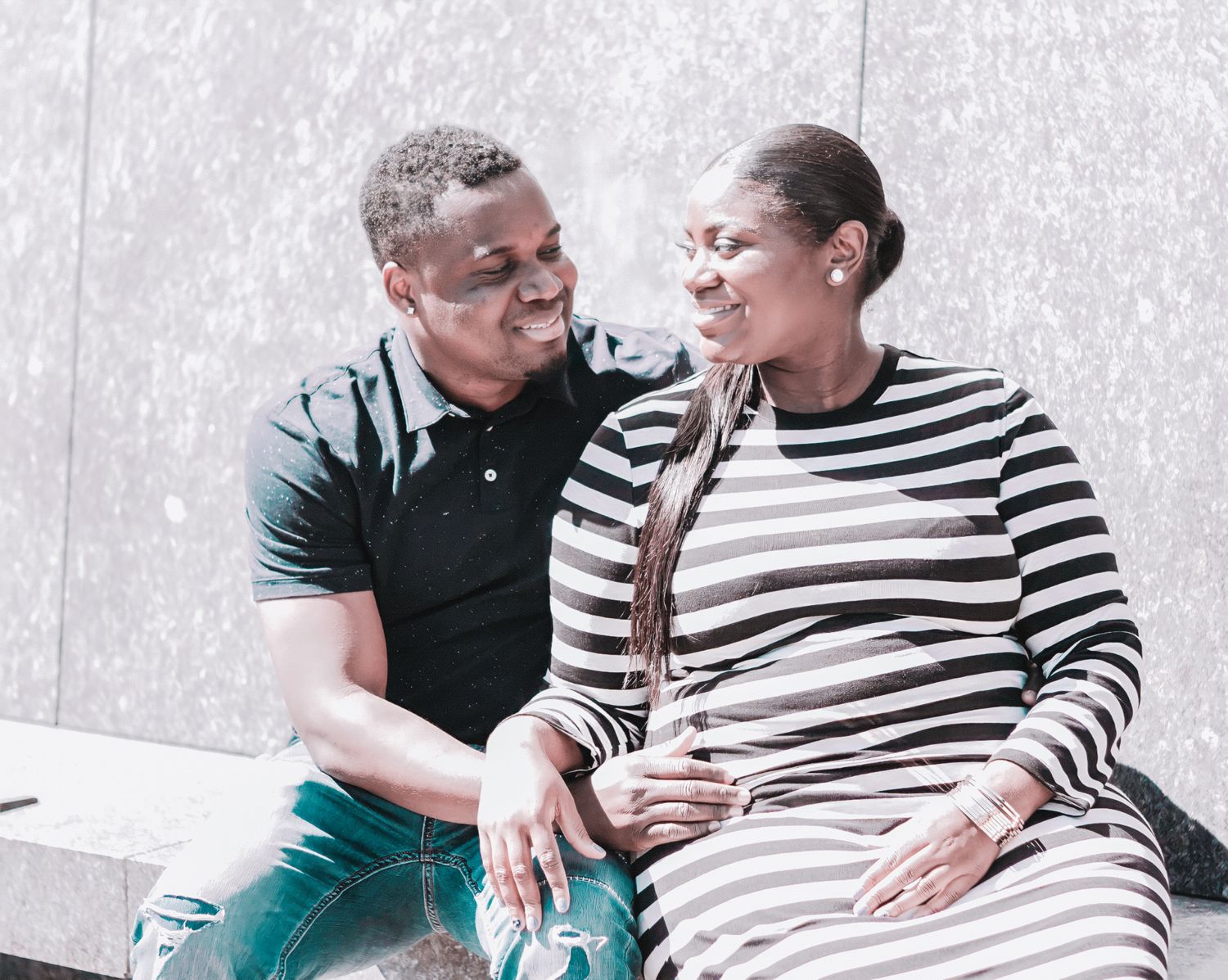 A smiling couple sits together on a bench, sharing a warm moment, the man in a polo shirt and woman in a striped dress.