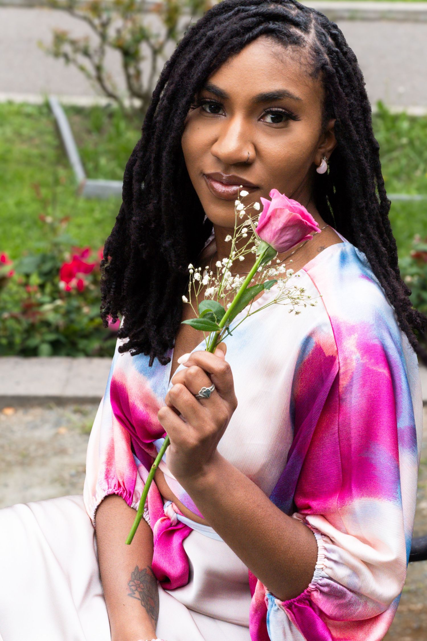 Elegant Black woman with locs in pink tie-dye top holding a single pink rose in a lush garden setting.