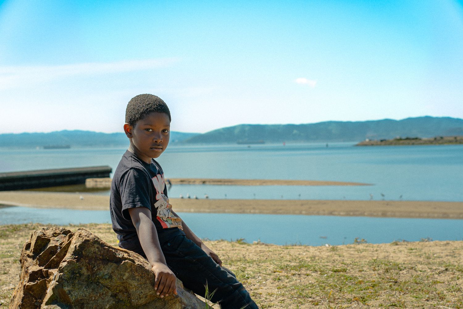 Young boy sitting on a rock by a scenic lake under a bright blue sky.