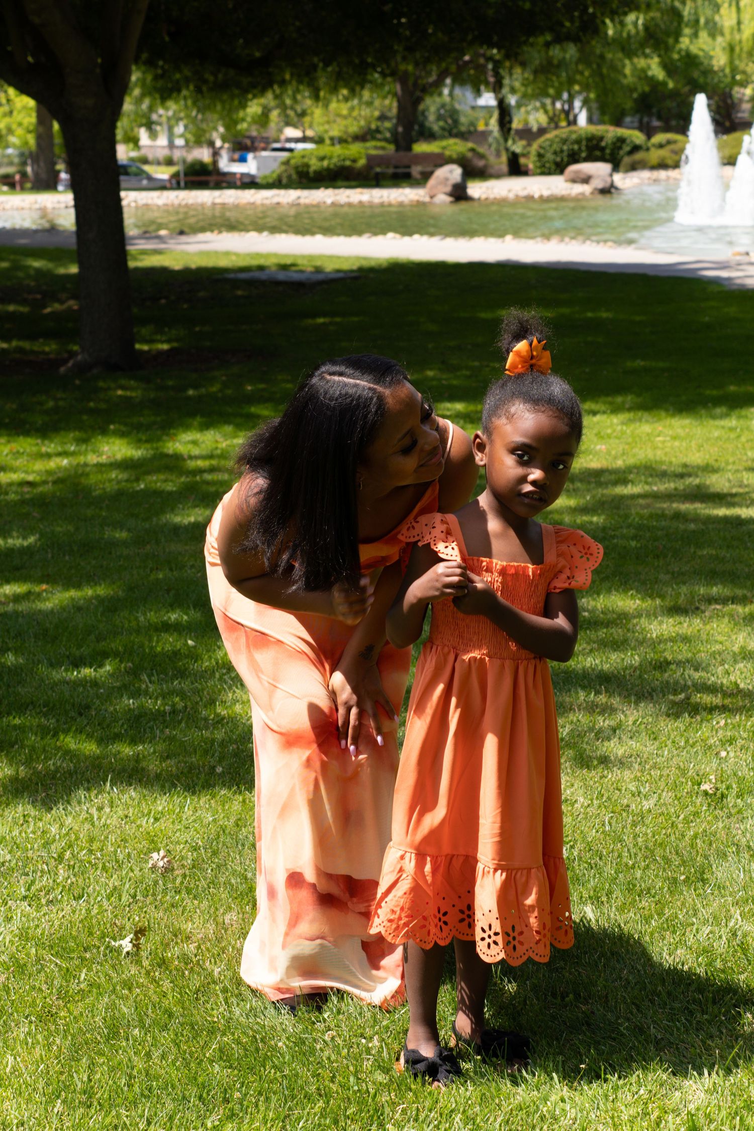 A Black woman in an orange dress kisses a young girl wearing a matching orange dress in a sunny park.
