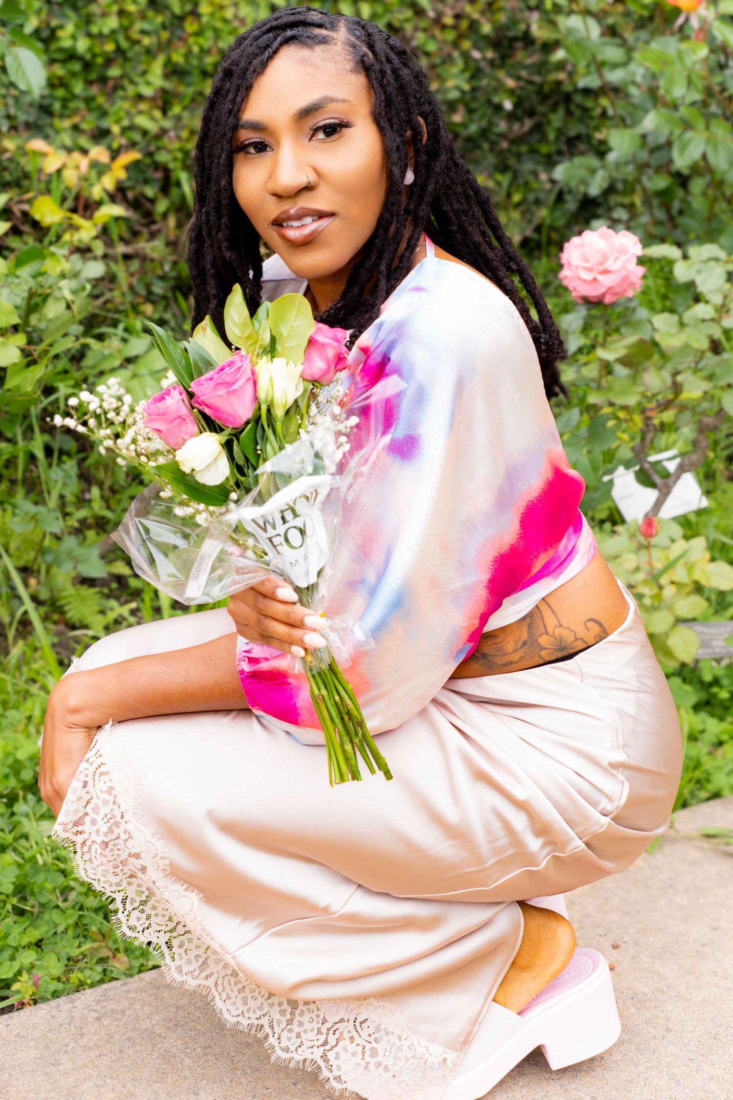 Woman in tie-dye top and satin skirt crouches holding pink rose bouquet in a garden.