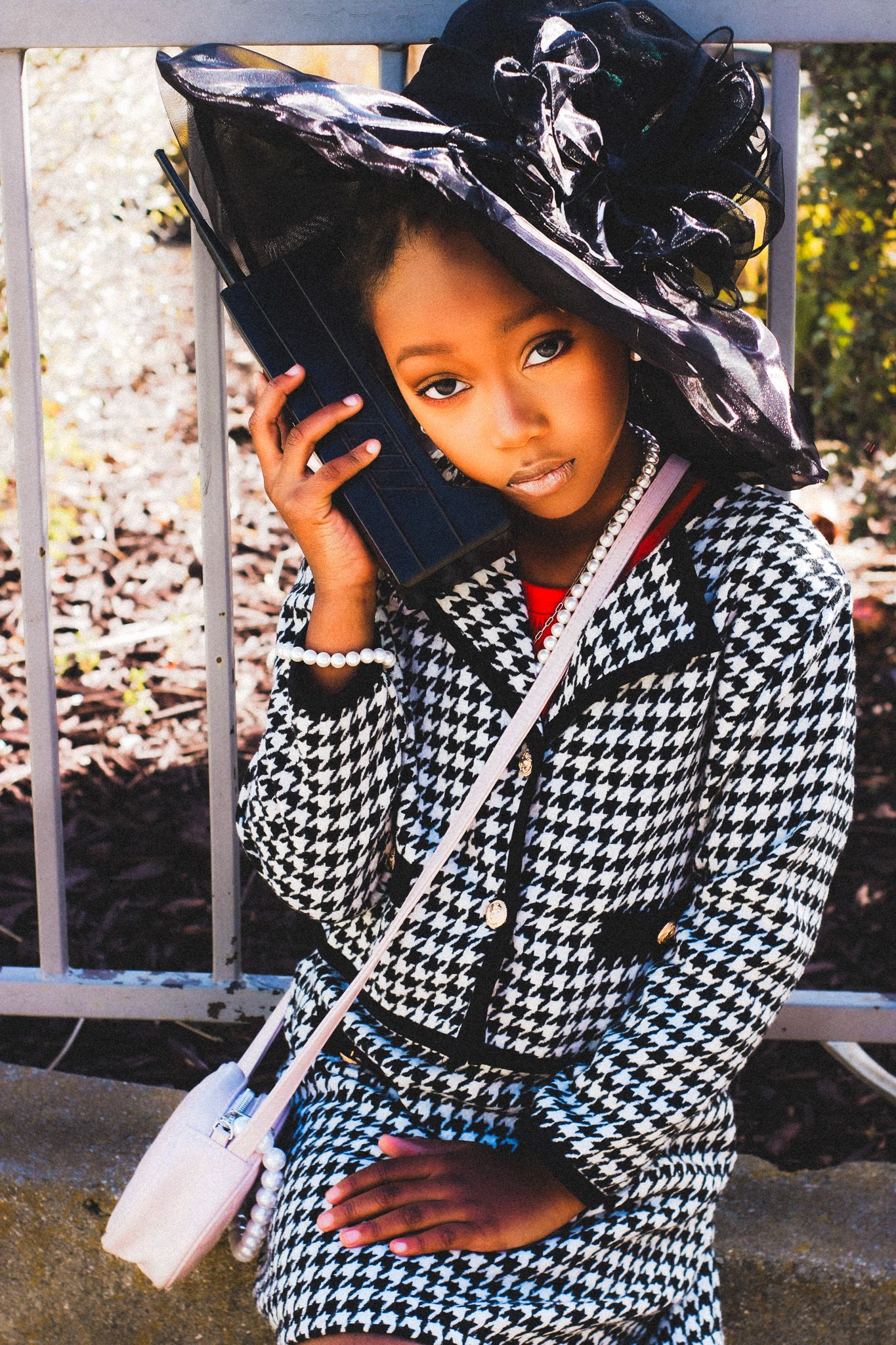 Stylish young Black girl wearing a houndstooth suit and black hat holding a phone outdoors.