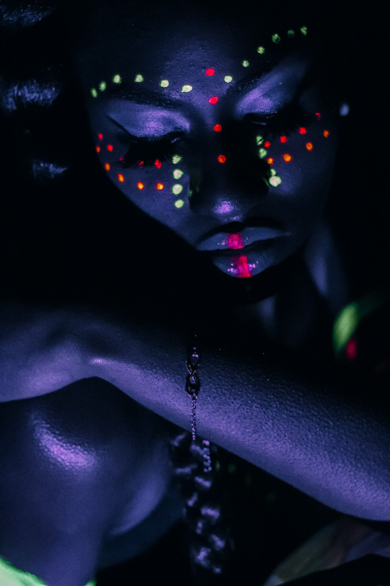 Woman with glowing neon UV body paint and colorful dot face art posing in dark blue light.