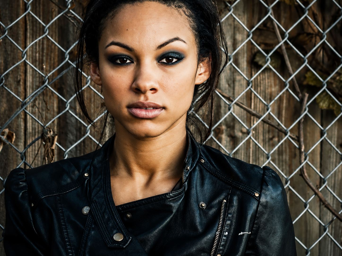 Young woman in black leather jacket poses confidently against chain-link fence with weathered wood background.