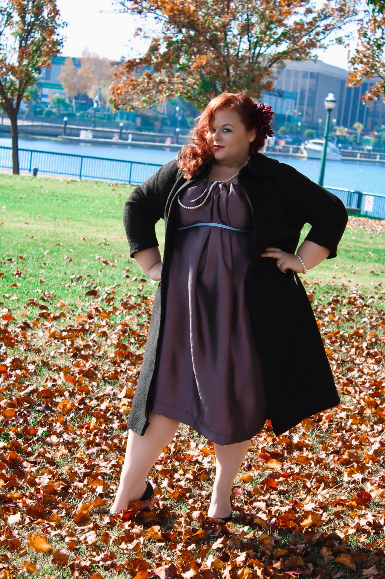Plus-size woman in purple dress and black cardigan poses among autumn leaves in a park by a river.