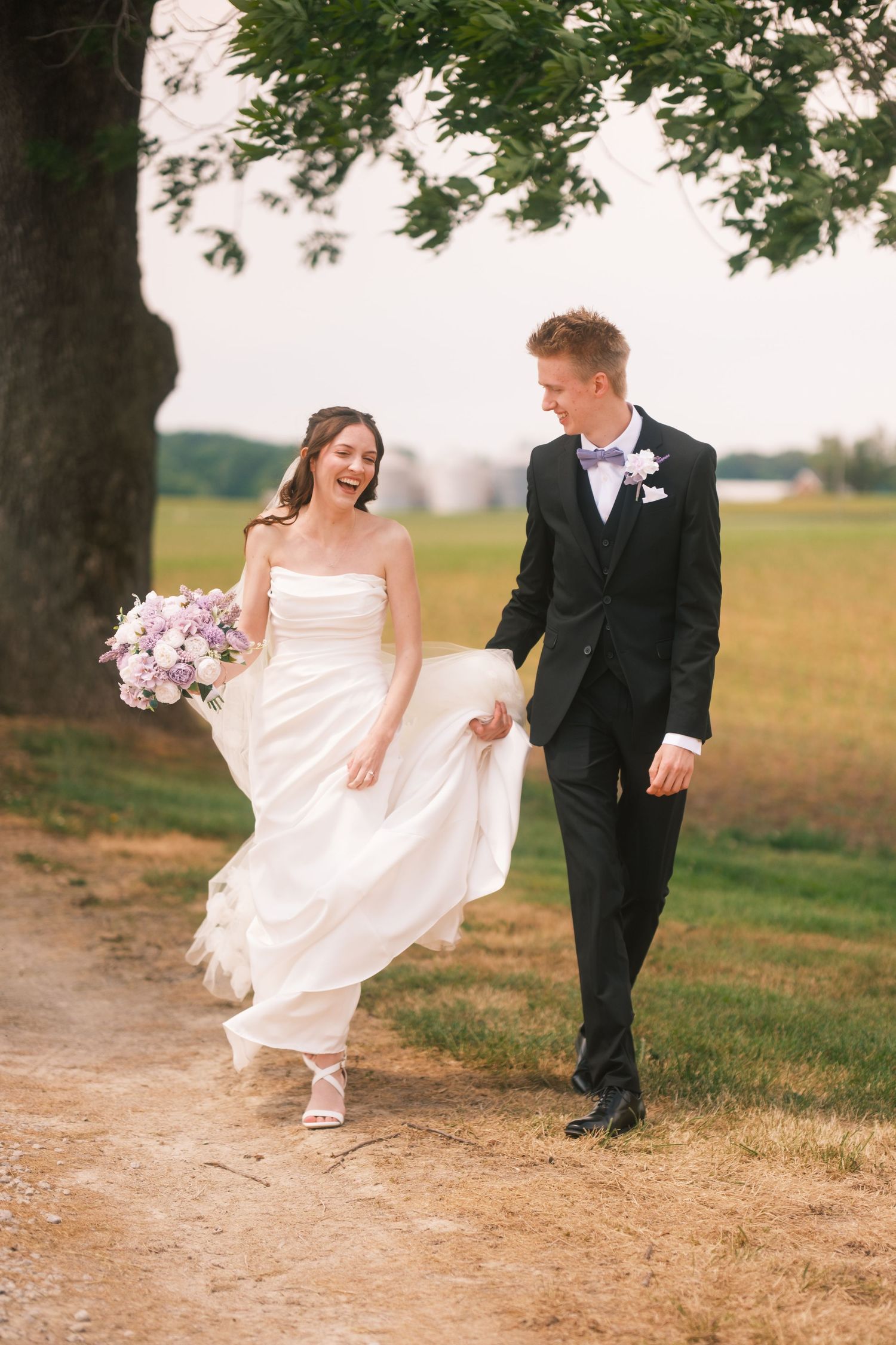 A joyful bride in a white gown holding purple flowers walks hand-in-hand with a groom in a black suit on a rural path.