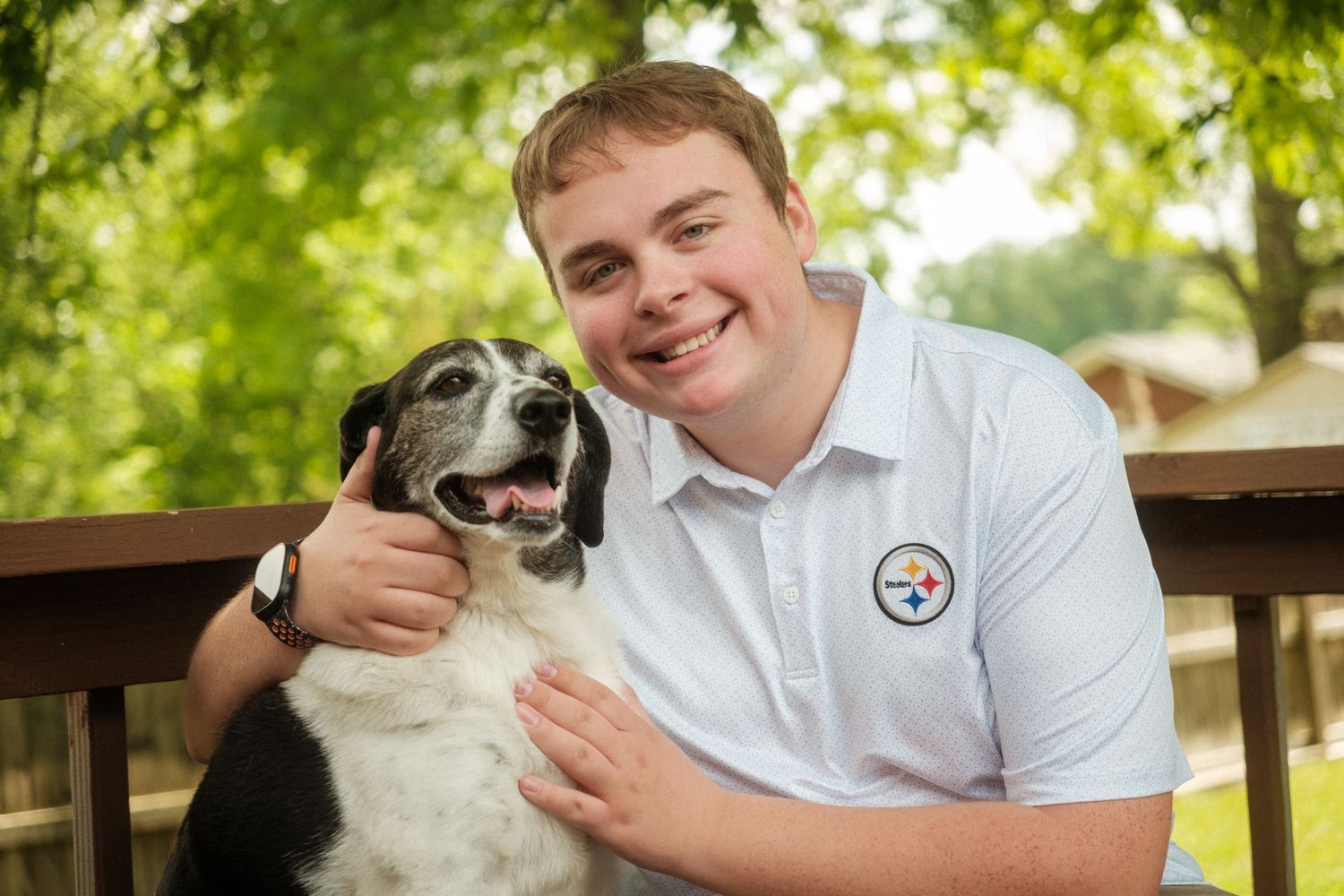 Smiling young man in Steelers shirt hugging a black and white dog on an outdoor deck surrounded by trees.