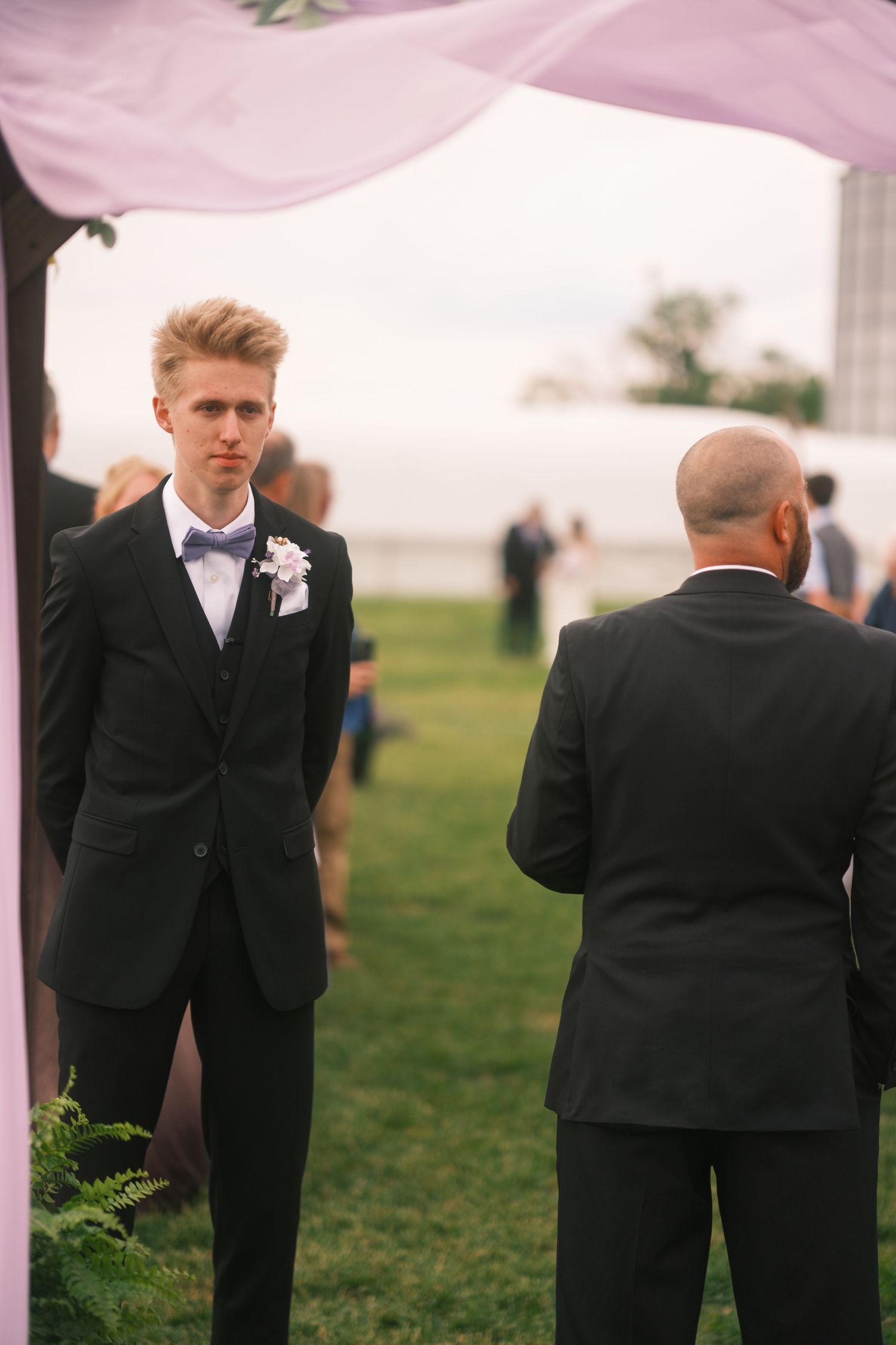 Young groom in black tuxedo with bow tie and boutonniere stands at outdoor wedding ceremony on green lawn.
