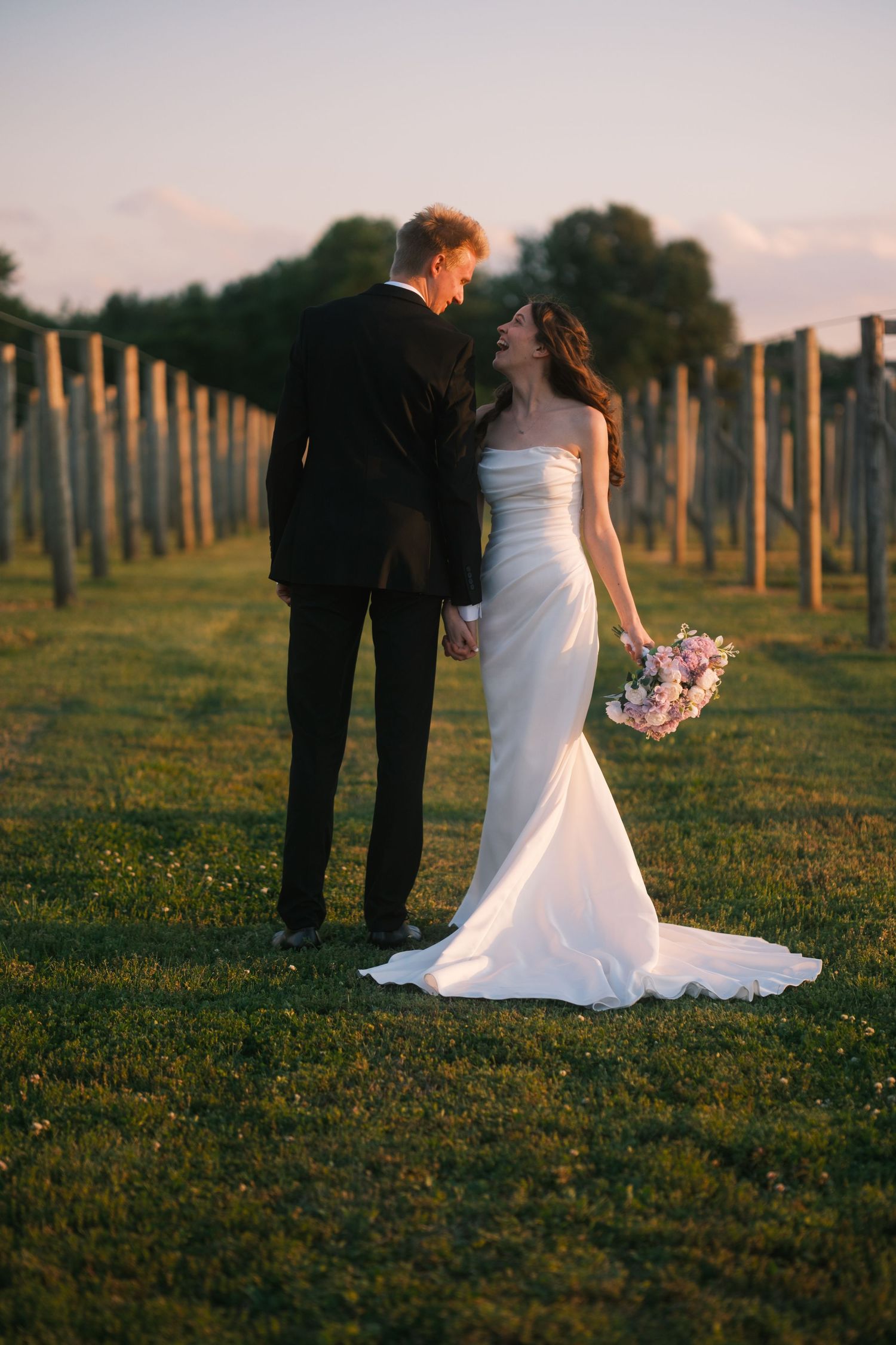 Bride in white gown laughing with groom in a vineyard at golden hour, holding pink bouquet.