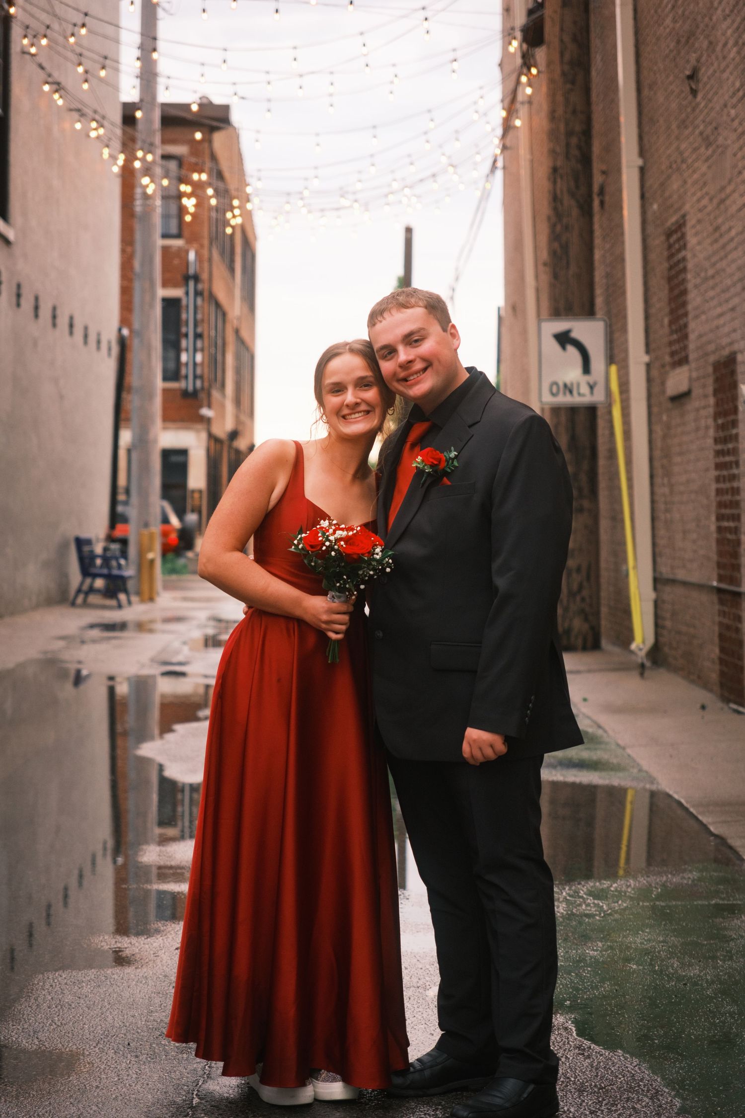 Couple in matching red dress and black suit posing in a rain-soaked alley with string lights overhead.