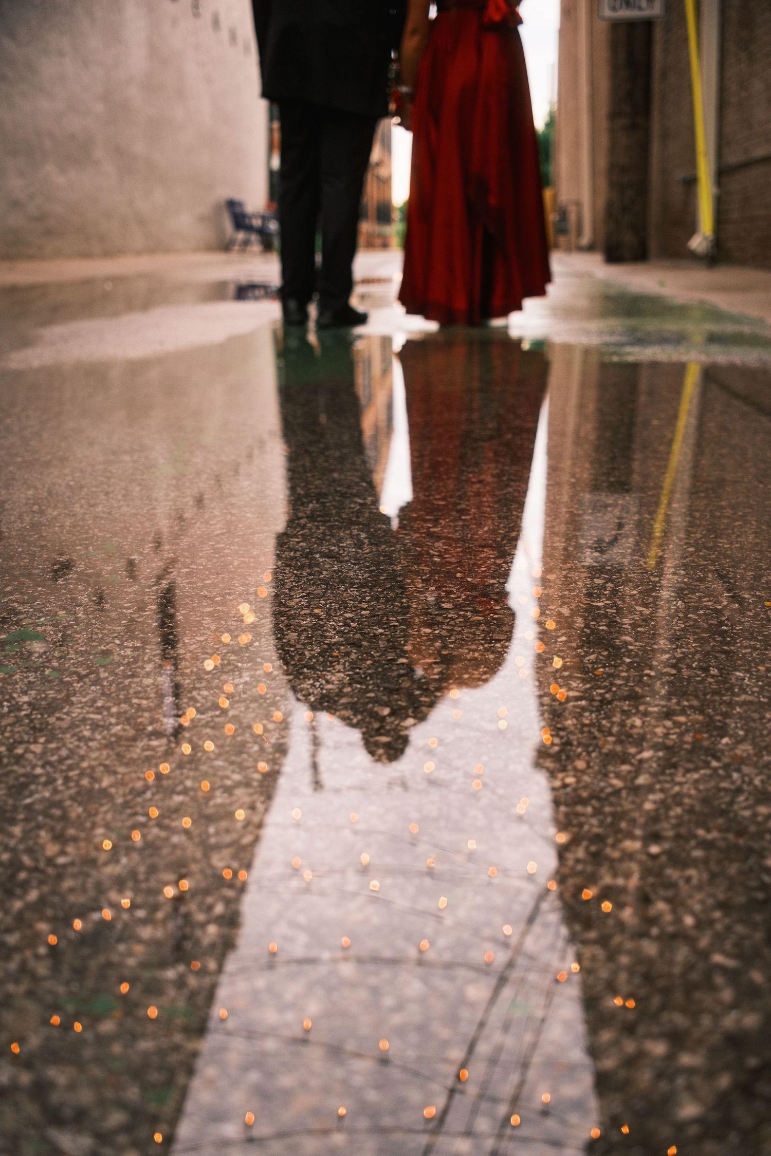 Reflection of couple in formal attire captured in rain puddle in a charming lit alleyway.