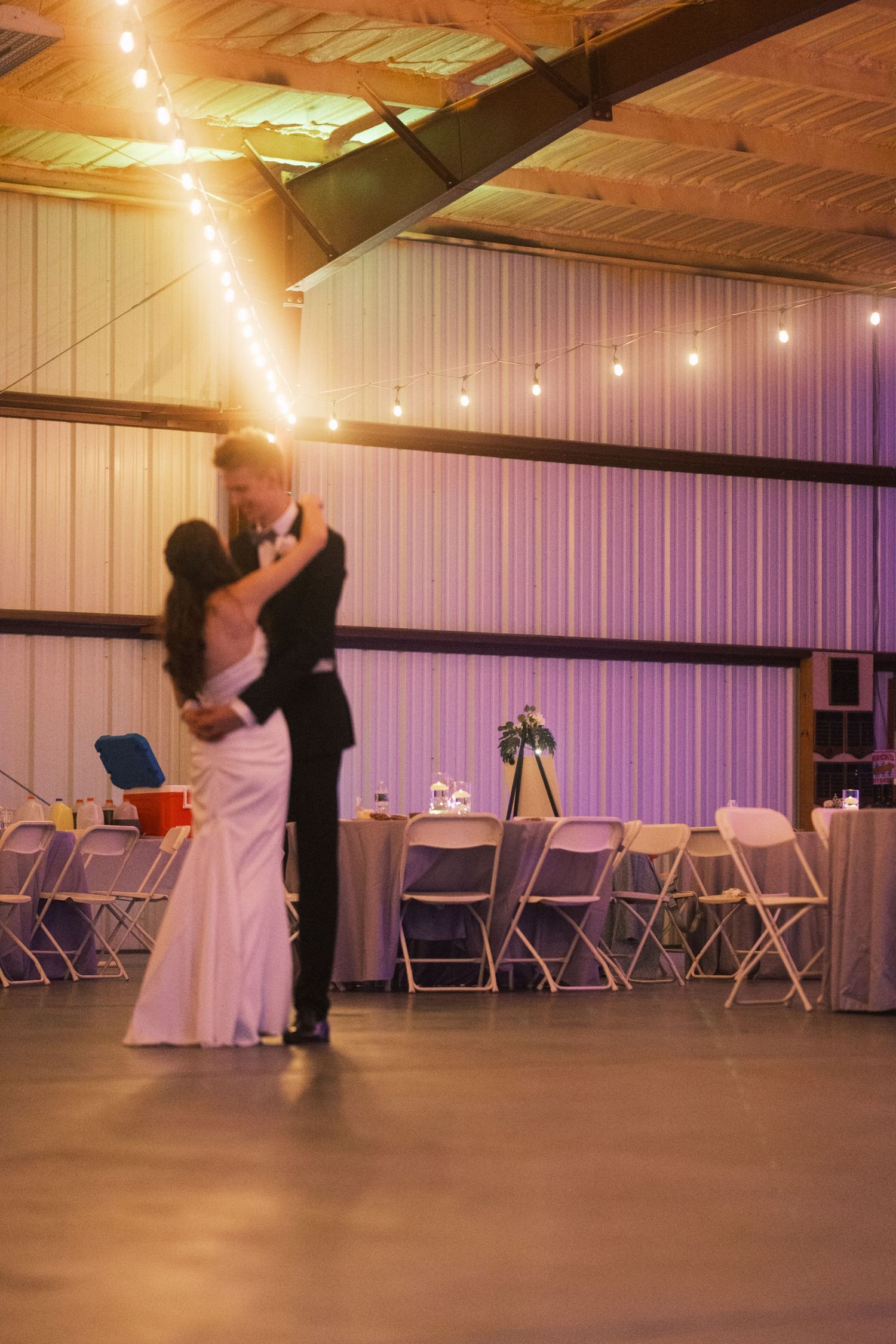 Bride and groom share their first dance in a rustic barn wedding venue with warm string lights overhead.