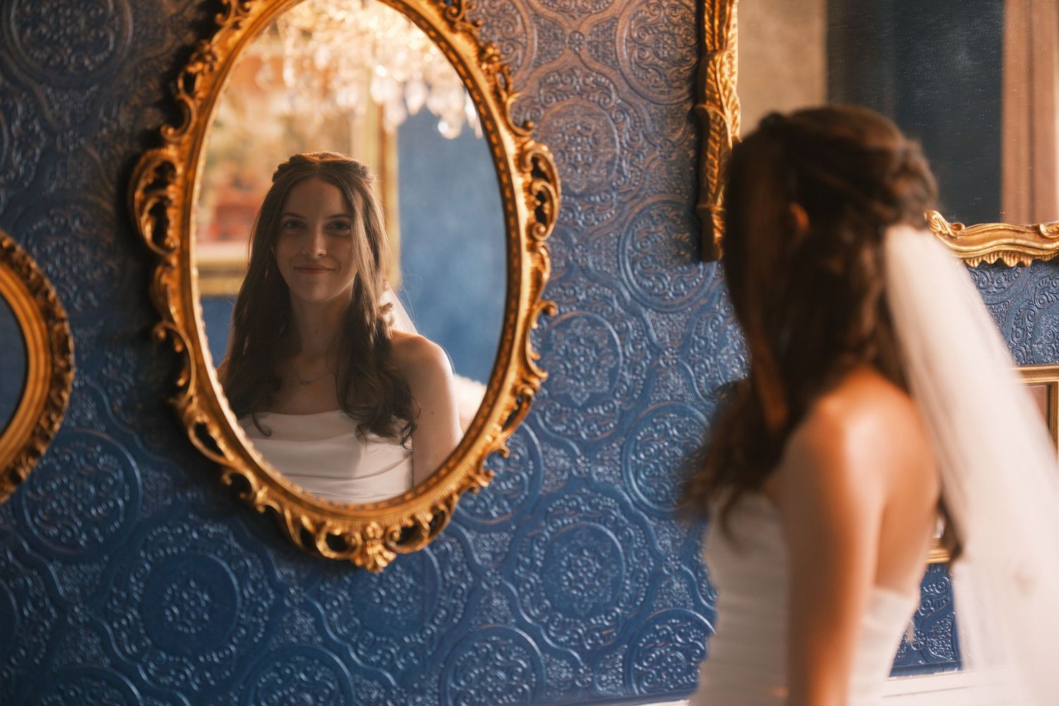 Bride in a white wedding dress admiring her reflection in an ornate gold mirror on a blue textured wall.