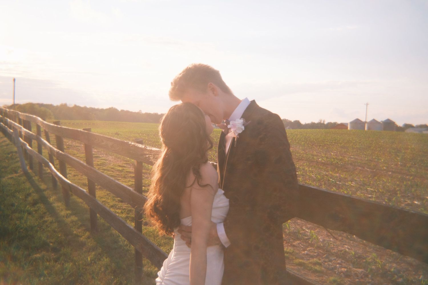 Bride and groom sharing a romantic kiss by a wooden fence at golden sunset.