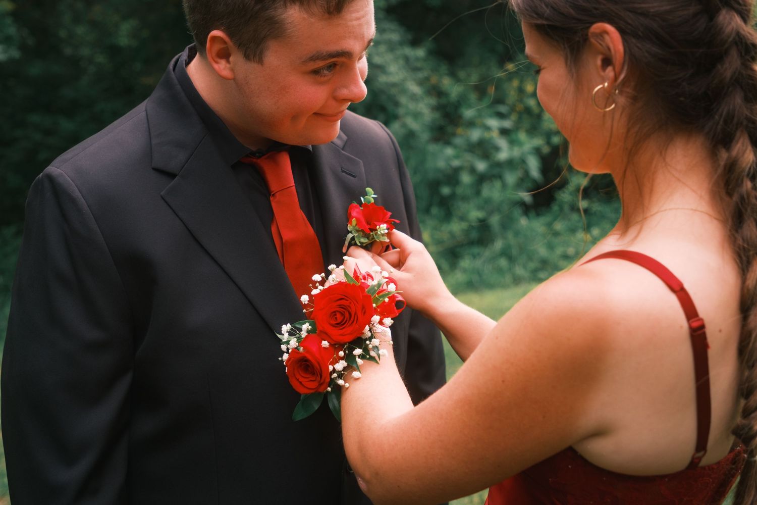 A young woman in a red dress pins a red rose boutonniere on a man in a black suit with red tie at prom.