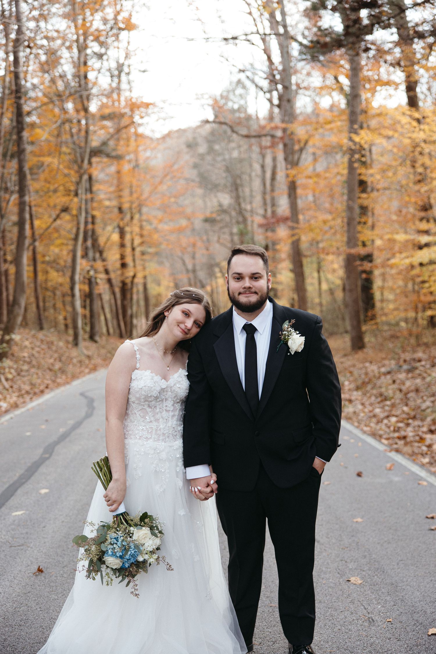 Bride and groom holding hands on a tree-lined road surrounded by vibrant autumn foliage during their fall wedding.
