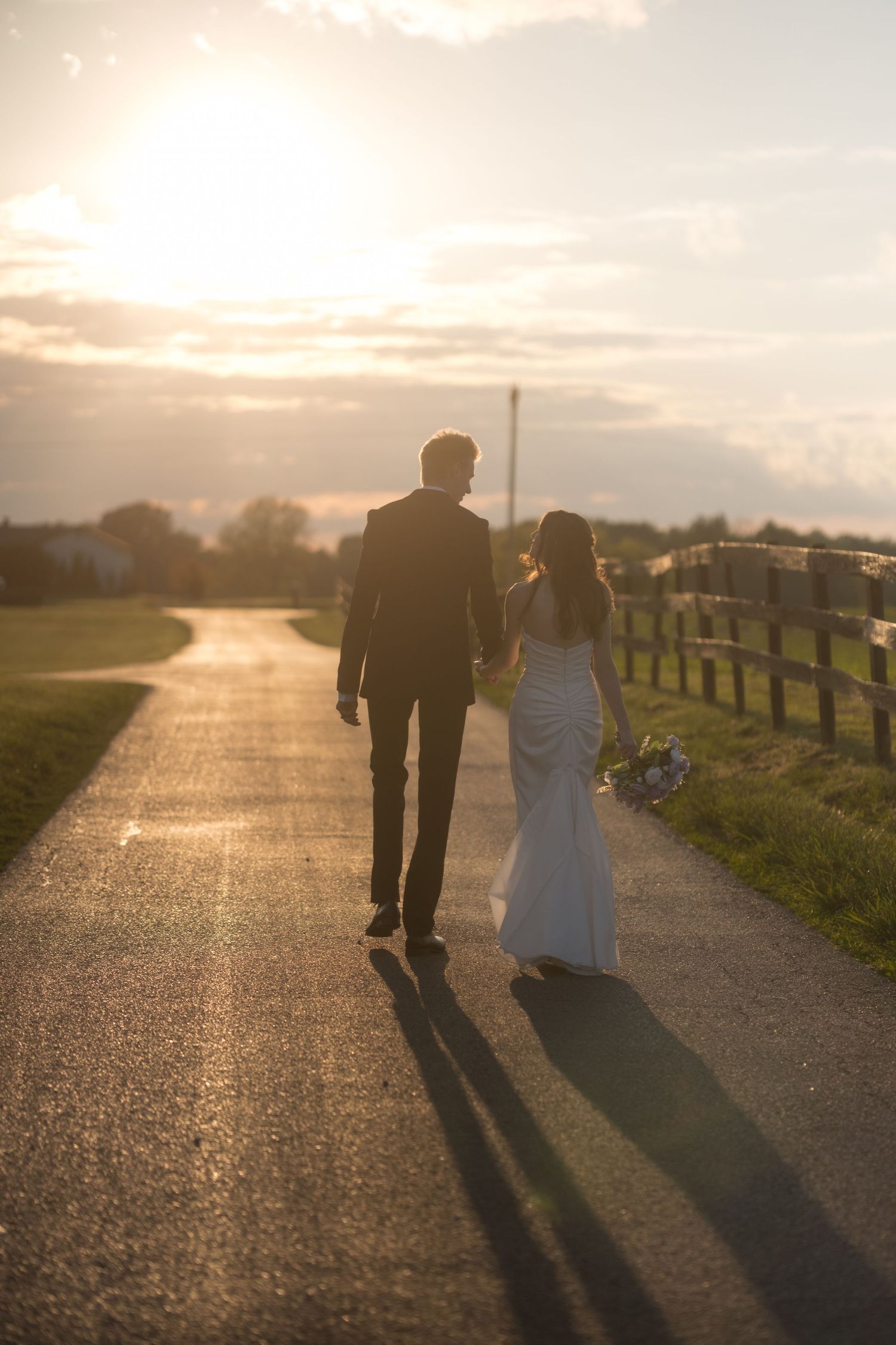 Newlyweds walking hand in hand down a country road at sunset, casting long shadows on the path.
