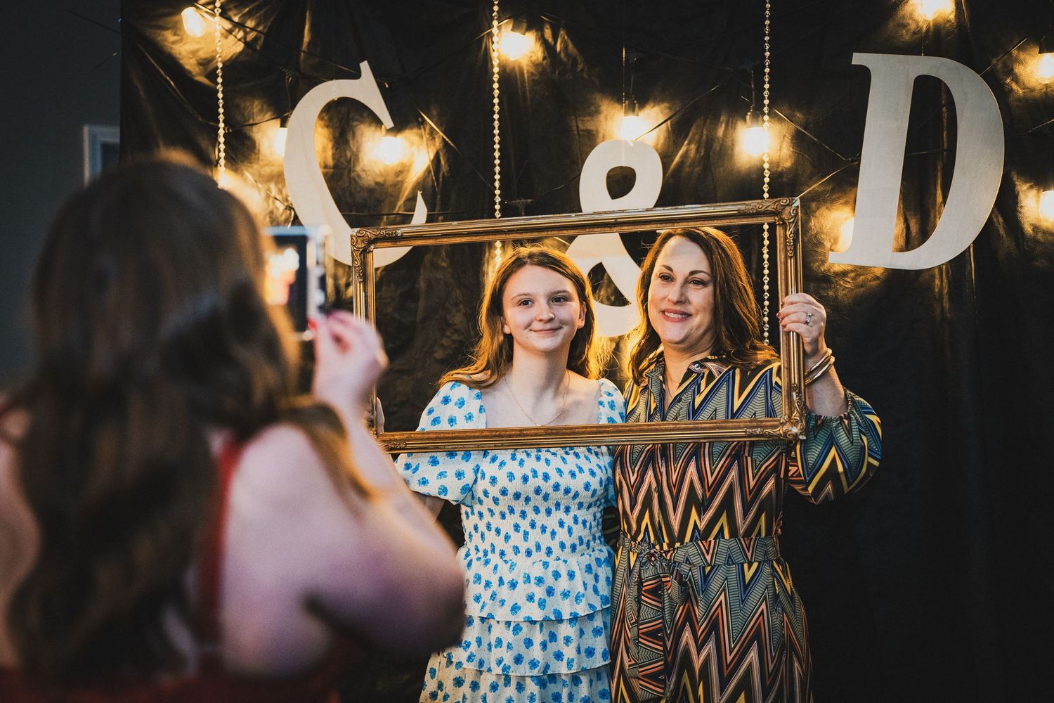Two women pose with a decorative gold frame at a party photo booth with string lights and monogram letters.