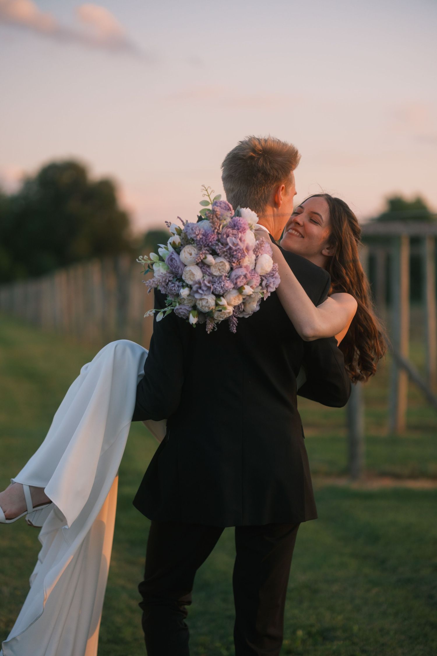 Groom carrying bride in black suit at sunset, she holds purple floral bouquet and smiles joyfully outdoors.