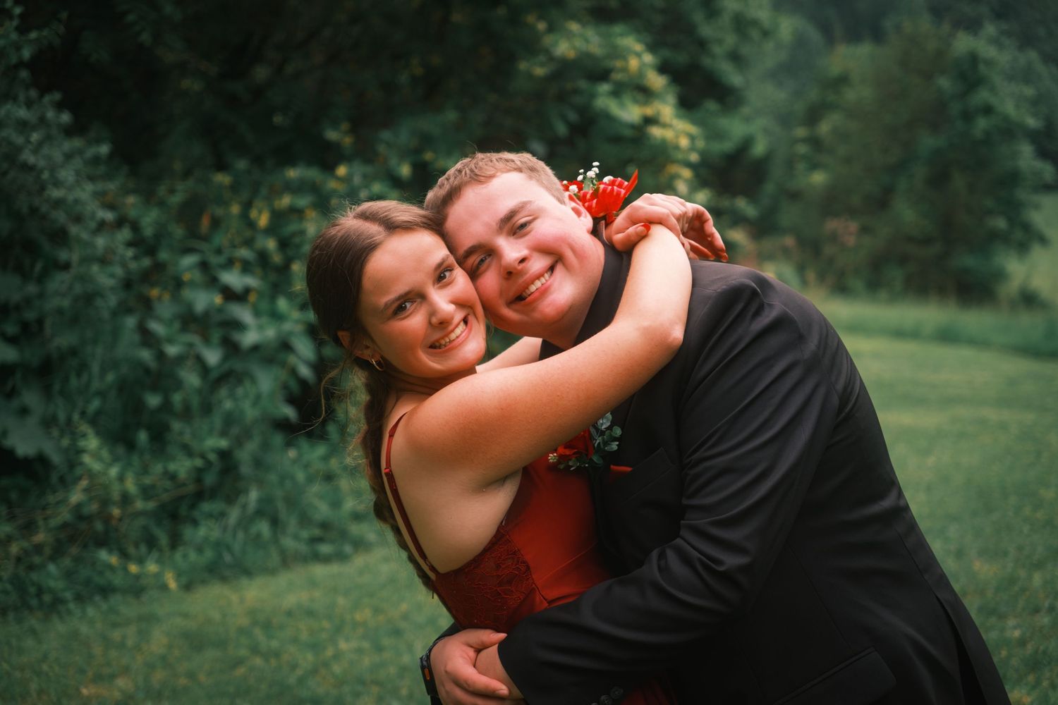 Happy prom couple hugging and laughing together in front of lush green outdoor backdrop.