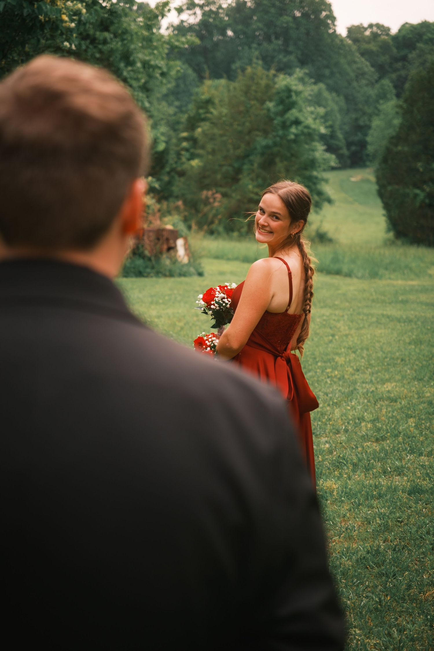 Smiling young woman in red prom dress looks back at her date on a green lawn.