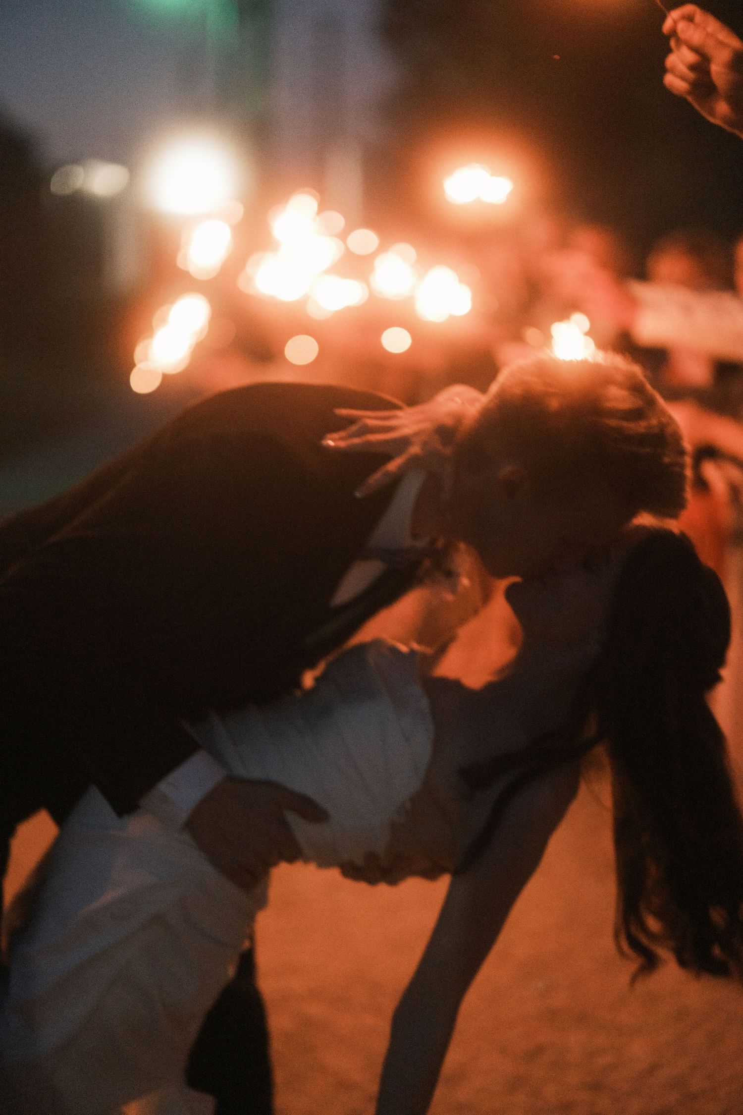 Couple sharing a romantic dip kiss surrounded by glowing sparklers at a nighttime wedding celebration.
