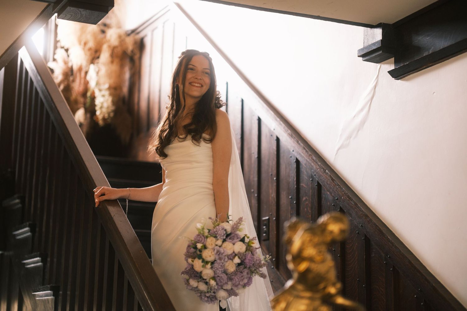Smiling bride in white gown holding purple bouquet descending elegant dark wood staircase.
