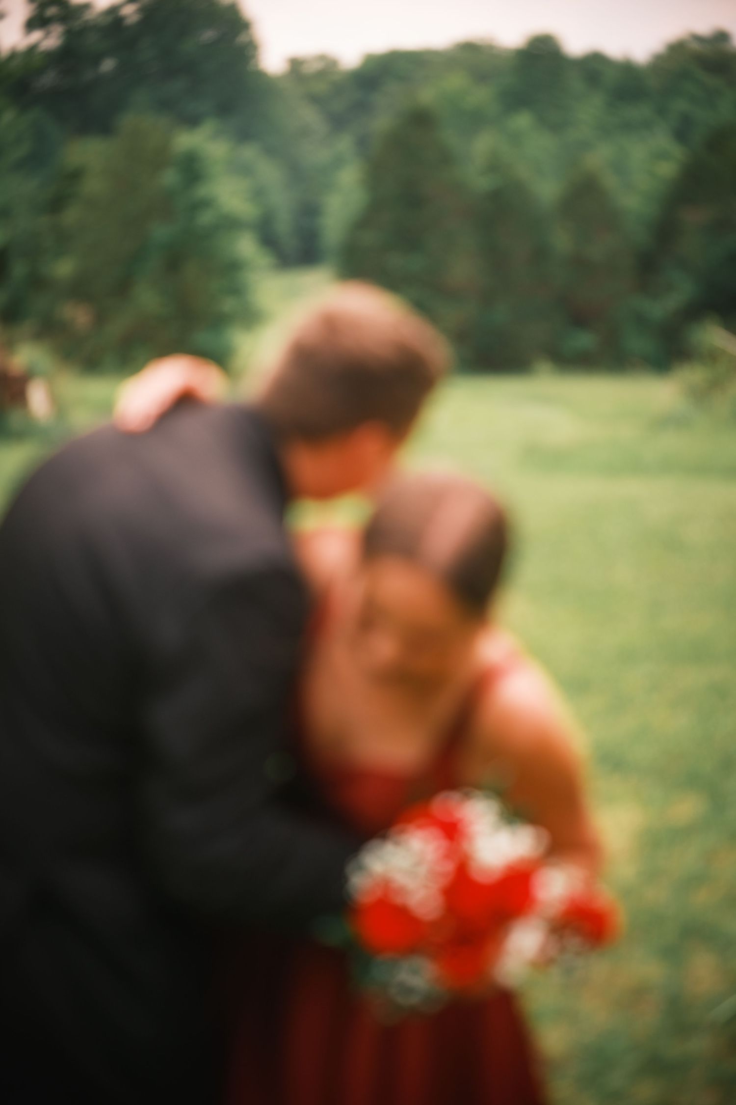 Blurry romantic couple embracing outdoors, woman in red dress holding bouquet, man in dark suit, green trees background.