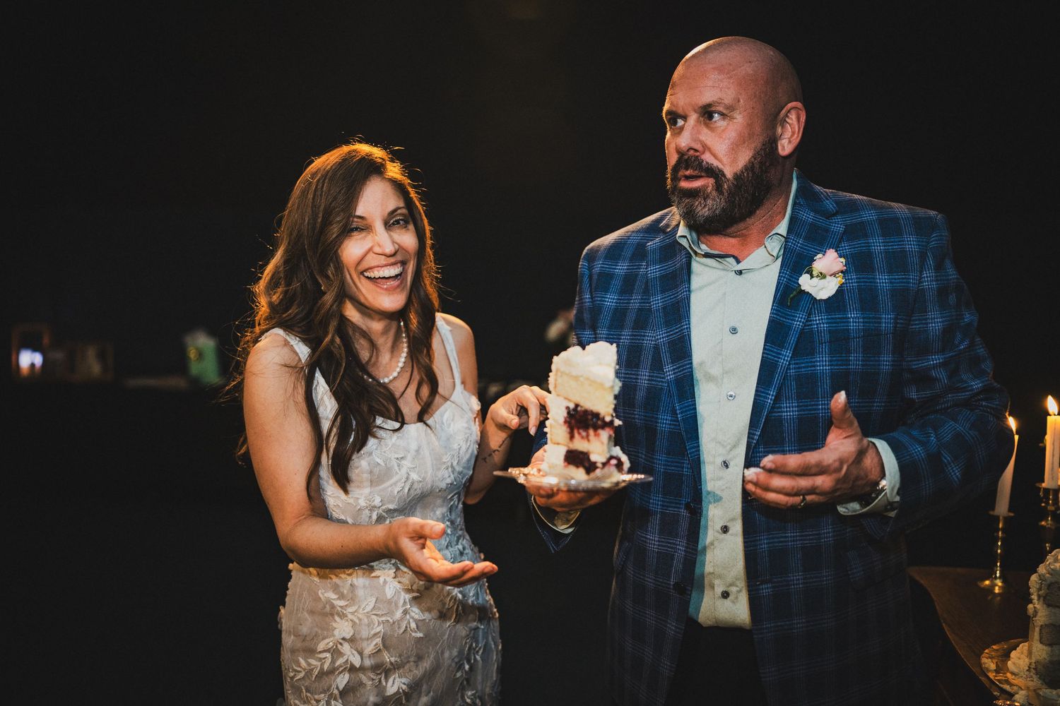 Bride and groom in blue plaid suit laughing while cutting wedding cake at reception.