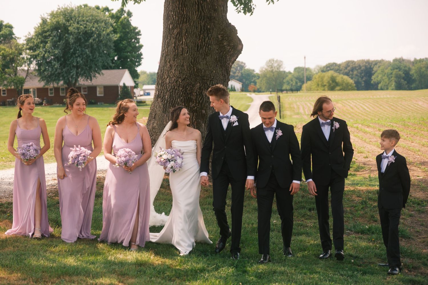 Wedding party poses outdoors near a tree, bridesmaids in lavender gowns and groomsmen in black tuxedos.