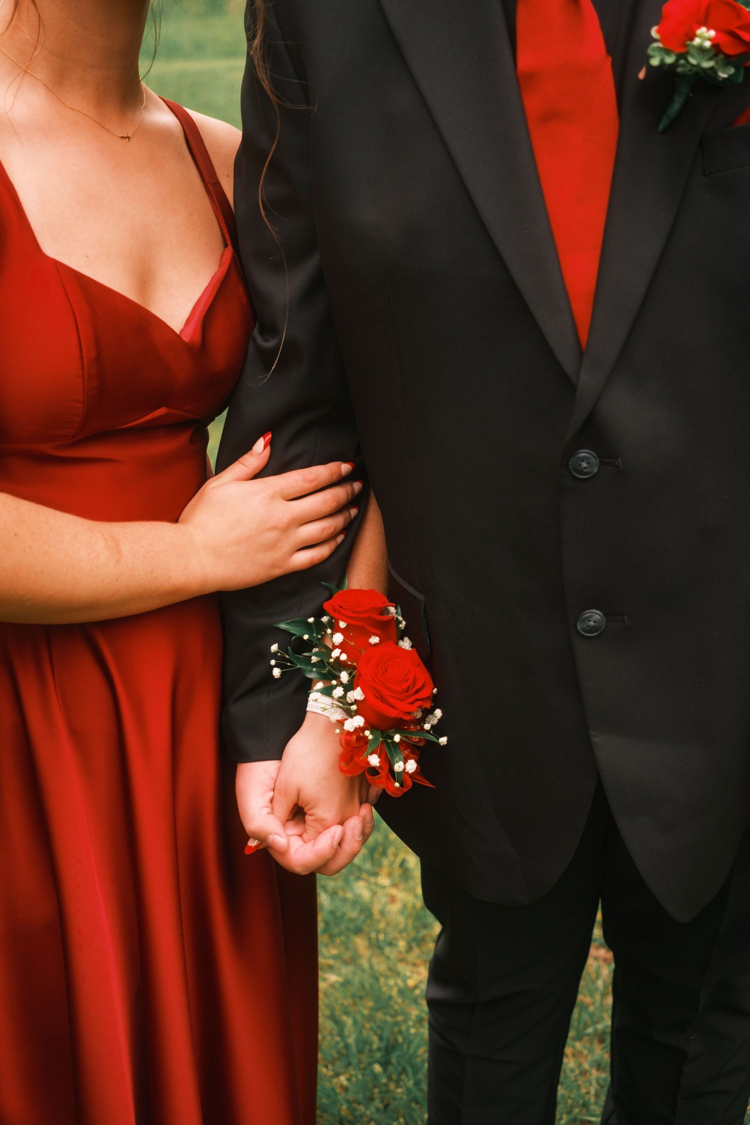 Couple in matching red and black prom attire, woman wearing red rose wrist corsage, holding hands outdoors.