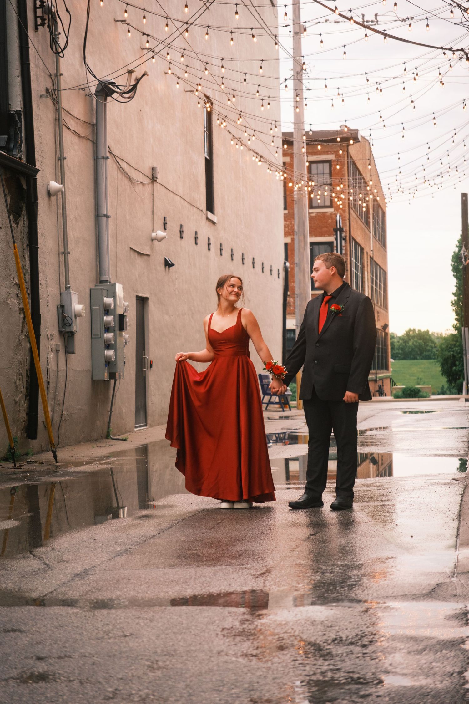 Couple in formal wear walks wet alley, woman in red gown, man in black suit, string lights overhead.