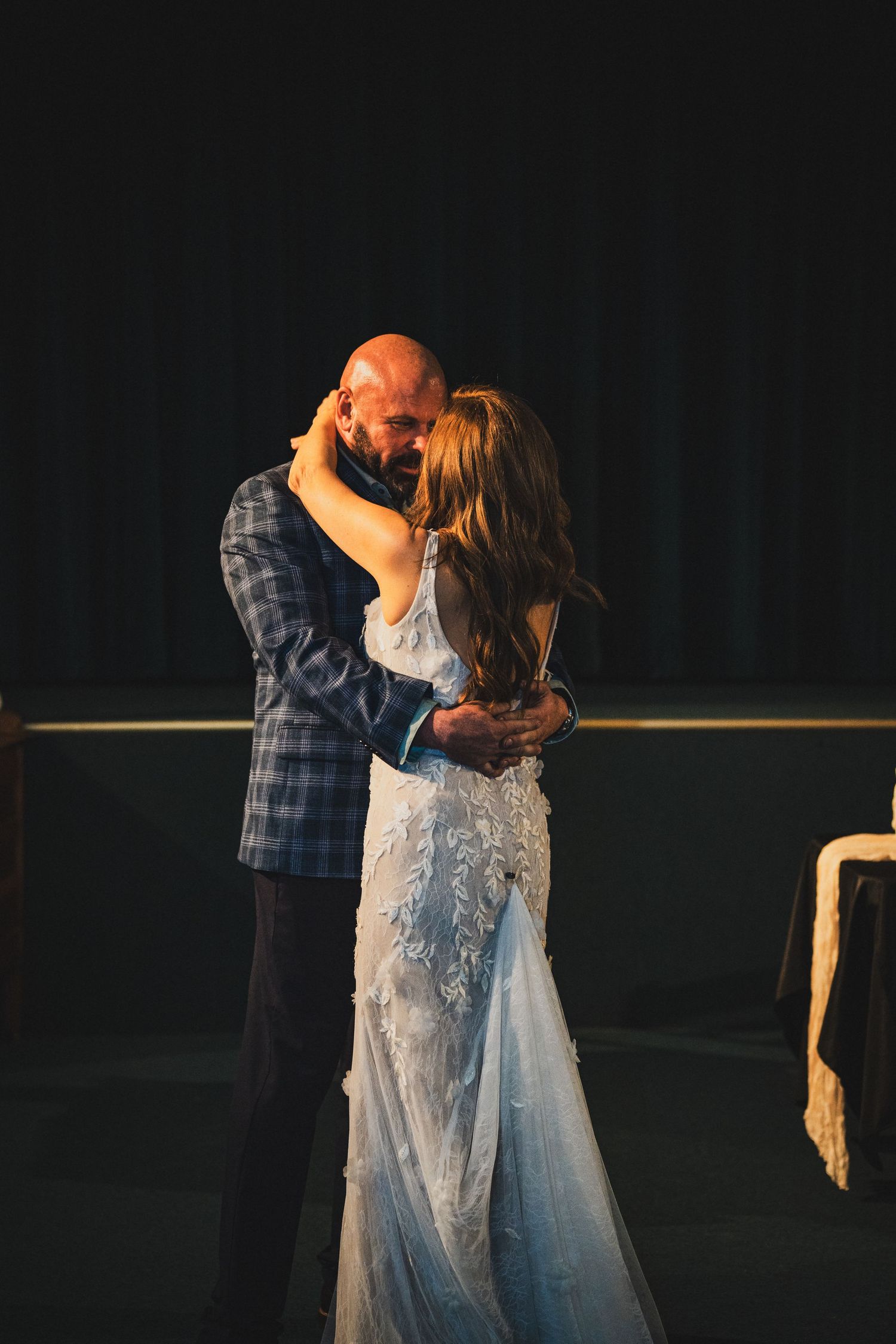 Bride in lace gown and bald groom sharing a romantic first dance in a softly lit venue.